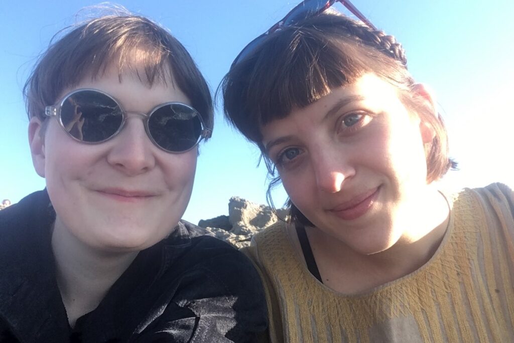 Two women lean in close to the camera, their faces almost touching. The woman on the left wears sunglasses with short cropped brown hair, the woman on the right with longer brown hair, her sunglasses perched on her head. Photo courtesy of artists