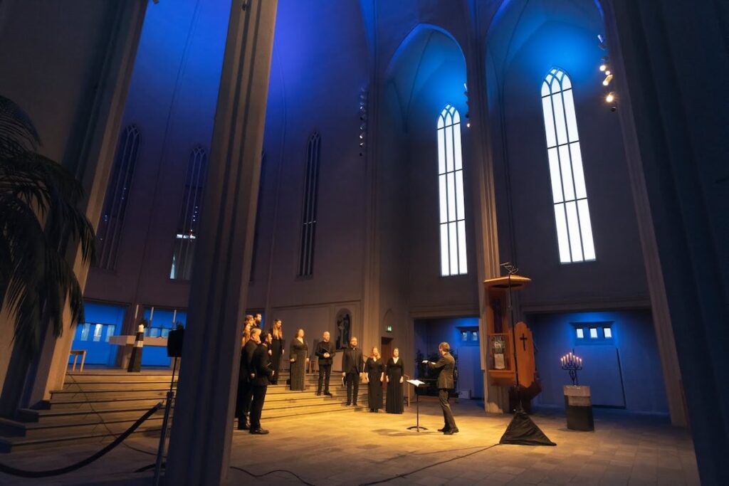 a choral group stands in a half circle in the sanctuary of an immense church, blue light emitting from the ceiling and the lower walls. Cantoque Ensemble, Dark Music Days 2026 -- Photo by Hans Vera