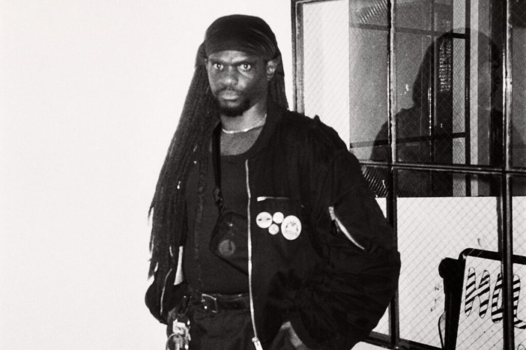 Image of Dreamcrusher, a dark-skinned African American man with long dreads, wearing a black sweatshirt  and standing in a front of a window. Black and white photo by Yaz Lancaster