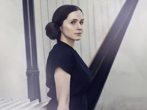 Image of a white woman with brown hair seated in front of a harp, body in profile, her face turned towards, wearing a short sleeve black shirt. Photo by Rut Sigurdardottir