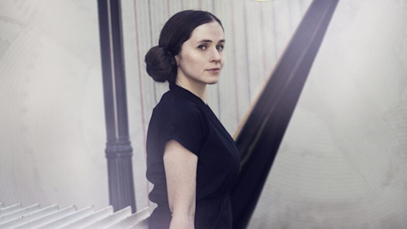 Image of a white woman with brown hair seated in front of a harp, body in profile, her face turned towards, wearing a short sleeve black shirt. Photo by Rut Sigurdardottir