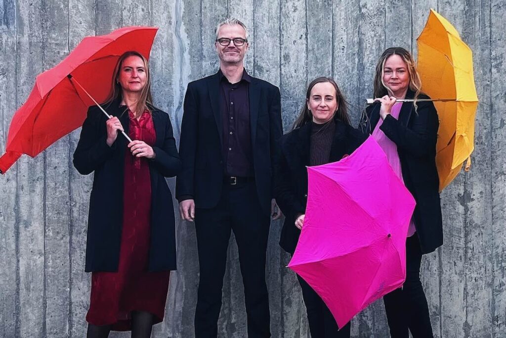 Four people stand in a row in front of a wall of grey wooden boards, a woman, a man, and two women; the women hold umbrellas, red, pink, and yellow, Dark Music Days 2026 -- Photo by Vala Halldors